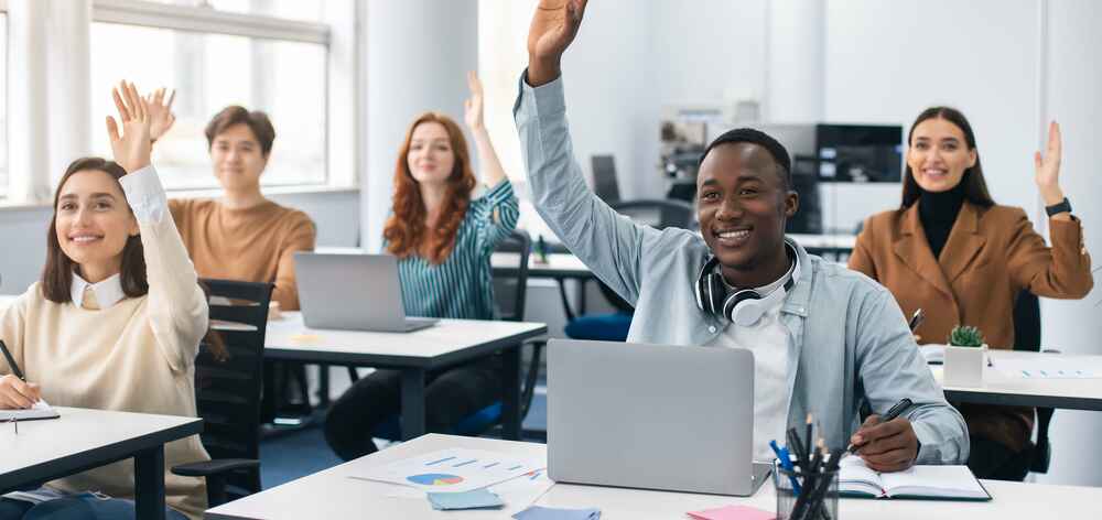 A group of students raising their hands in a classroom with laptops on their desks.