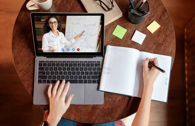 Above top view of female student sitting at desk, writing in notebook, taking notes, using laptop, having online class with teacher.