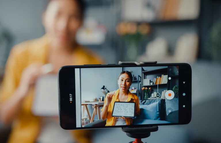 A woman in a yellow shirt holding a whiteboard is being recorded by a smartphone on a tripod for a video.