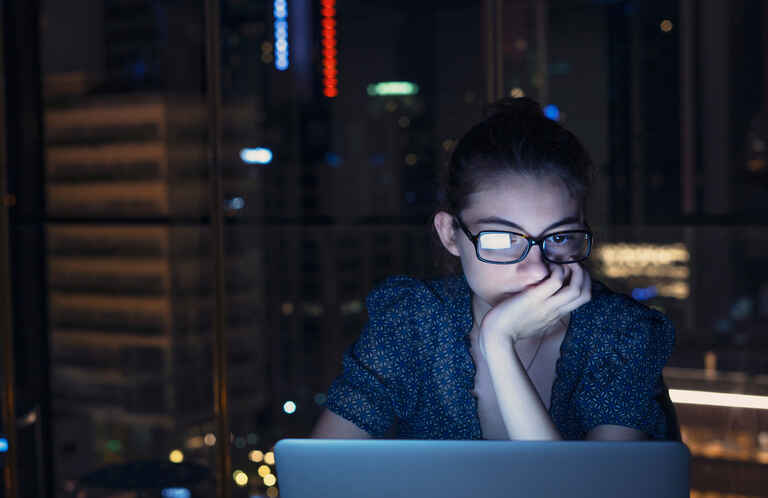 serious woman is highlighted by the glow of a computer screen in front of a city at night time