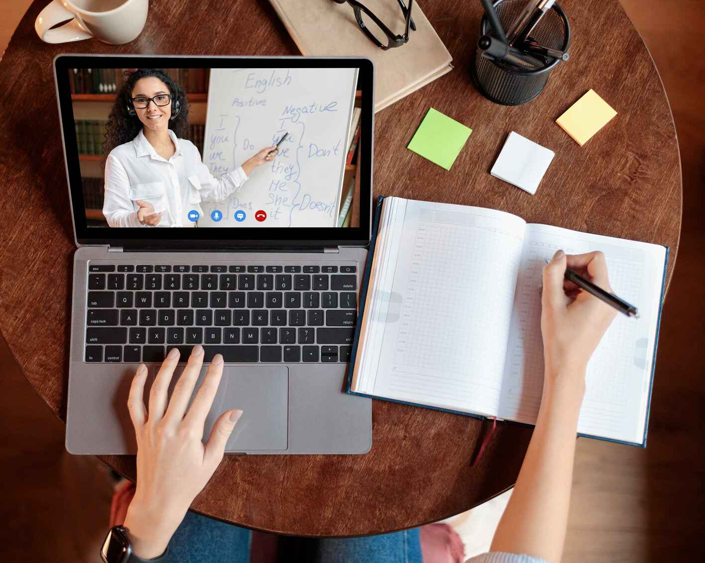 Above top view of female student sitting at desk, writing in notebook, taking notes, using laptop, having online class with teacher.