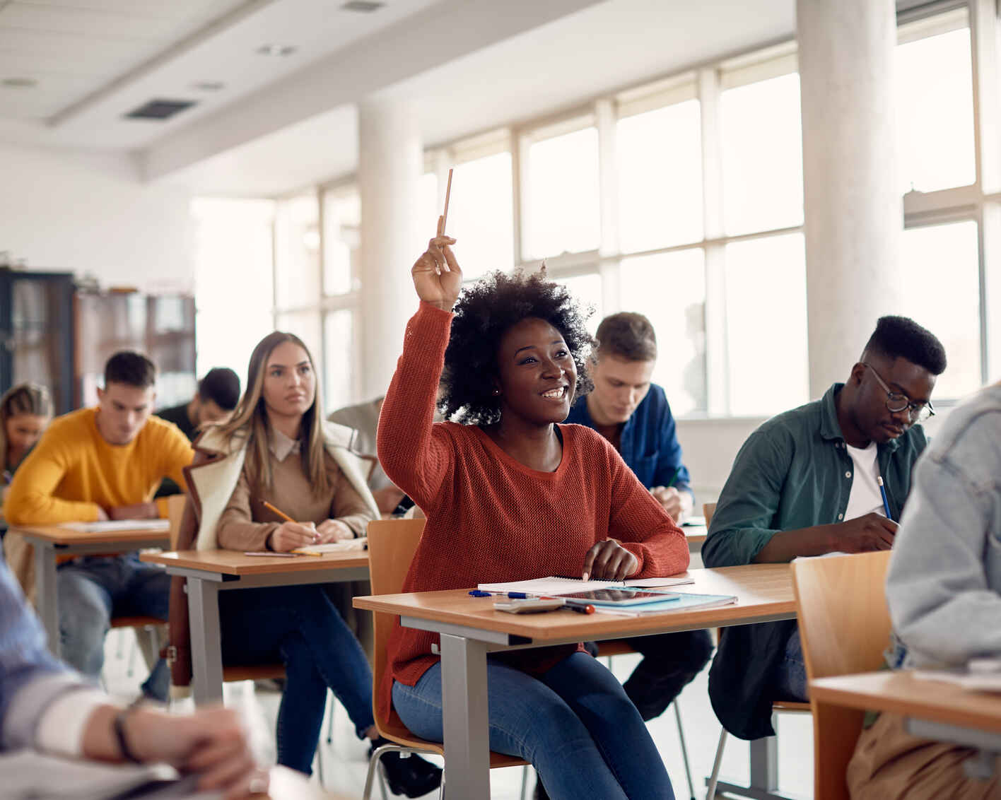 Happy black student raising arm to answer question while attending class with her university colleagues.