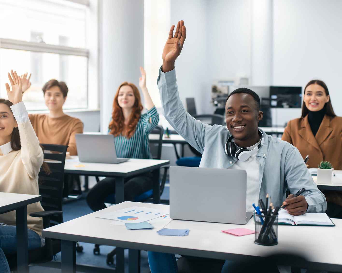 A group of students raising their hands in a classroom with laptops on their desks.