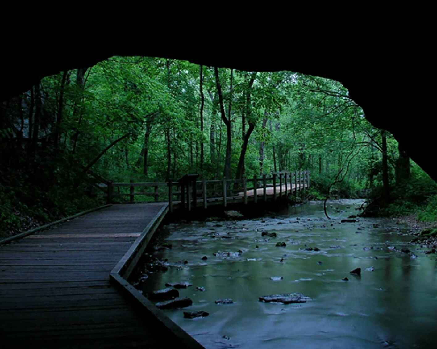 View from a cave opening onto a serene wooden boardwalk next to a river in a lush, green forest.