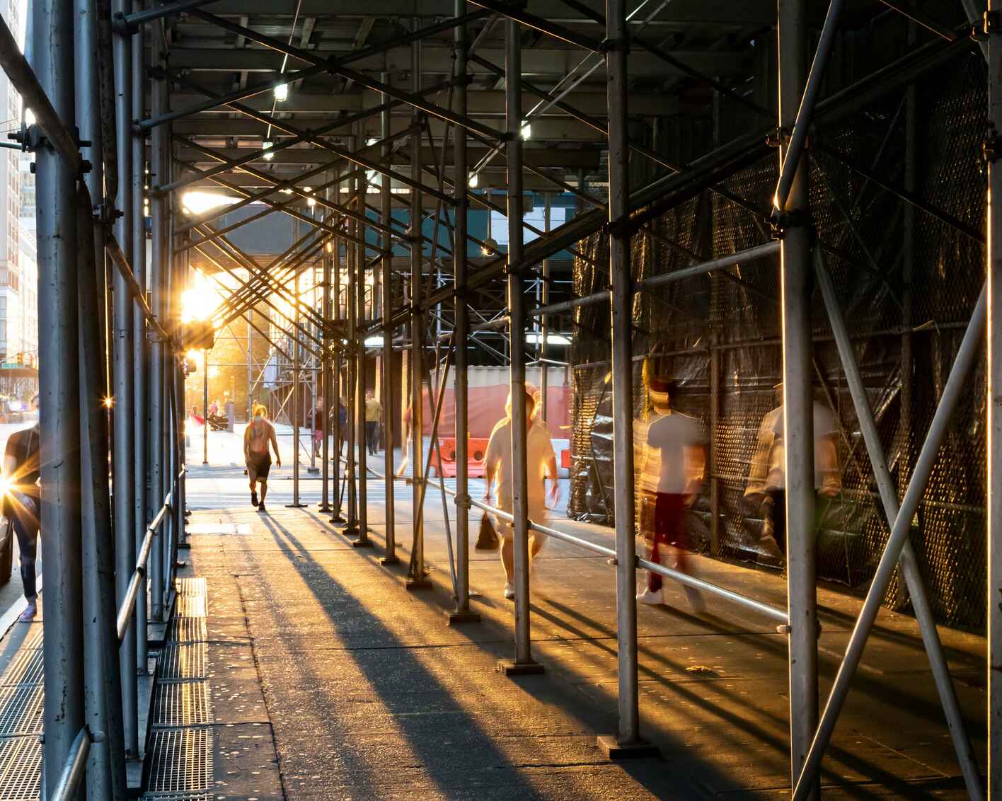Sunset urban scene with people walking under construction scaffolding on a city sidewalk, casting long shadows. Cars and buildings are visible in the background.