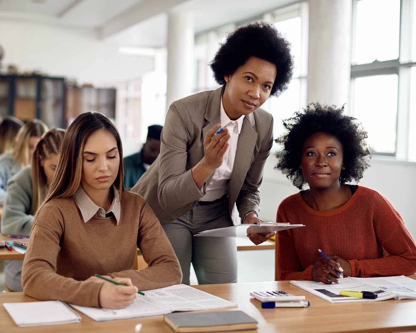 A teacher assists students in a classroom, one student looking up attentively while another focuses on her work, with more students studying in the background.