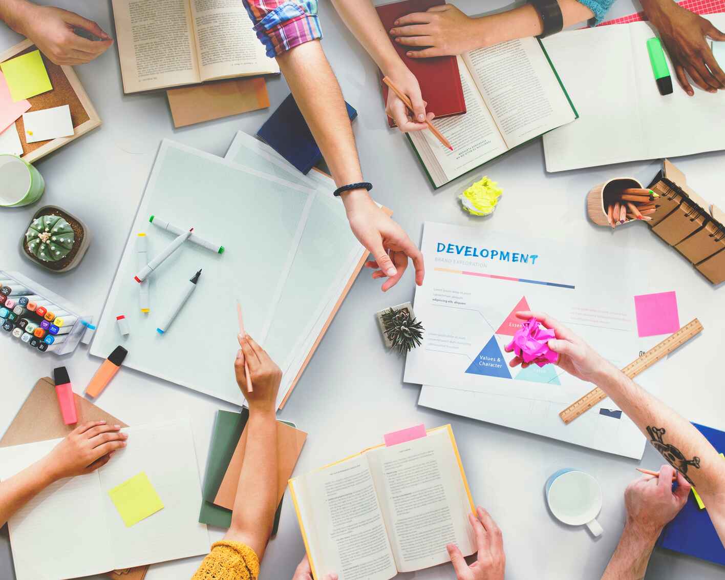 A group of people working together at a table filled with books, notebooks, markers, sticky notes, and a development project sheet, indicating a collaborative brainstorming session.