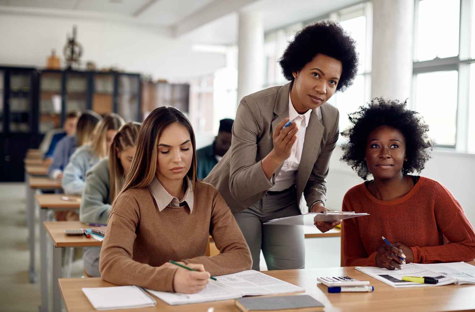 A teacher assists students in a classroom, one student looking up attentively while another focuses on her work, with more students studying in the background.