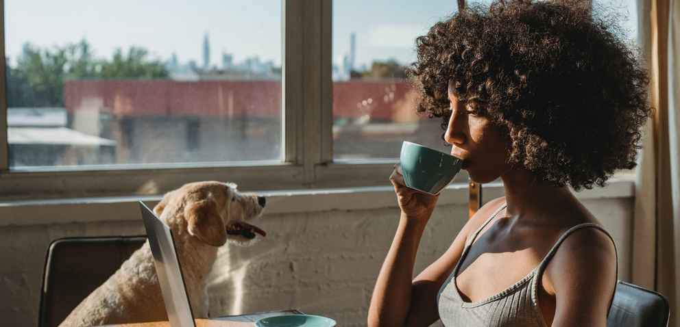 woman of color sipping coffee while browsing on her laptop and her dog sits close by