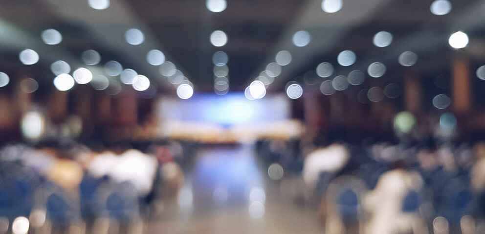 Blurred image of a large room filled with chairs facing a stage.