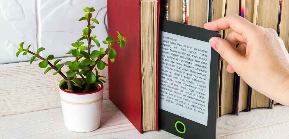 Woman hand takes e-reader from shelf with paper books and small potted plant