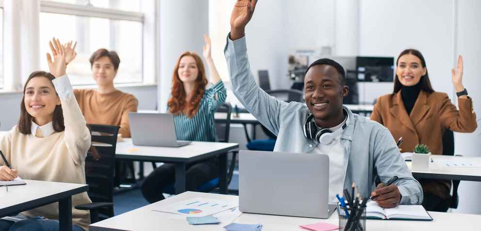A group of students raising their hands in a classroom with laptops on their desks.