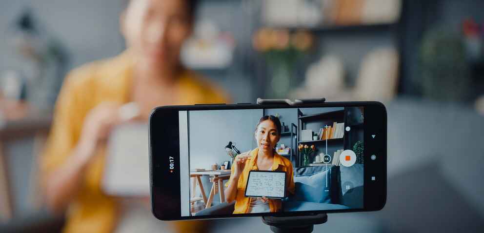 A woman in a yellow shirt holding a whiteboard is being recorded by a smartphone on a tripod for a video.
