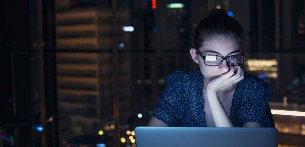 serious woman is highlighted by the glow of a computer screen in front of a city at night time
