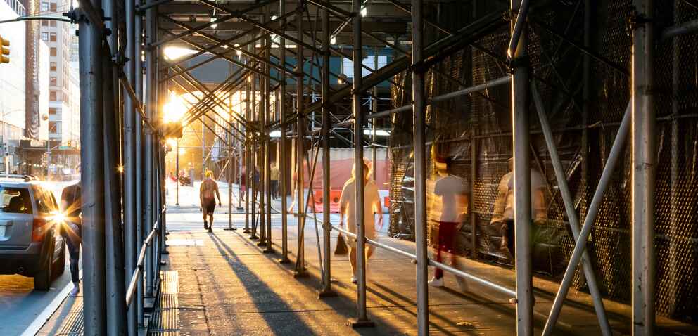 Sunset urban scene with people walking under construction scaffolding on a city sidewalk, casting long shadows. Cars and buildings are visible in the background.