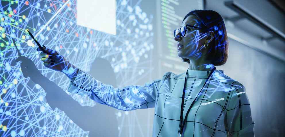 A woman in glasses points to a large screen displaying a complex network of interconnected nodes, illuminated by blue and white light patterns.