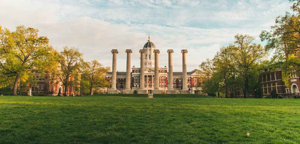 A grassy quad with six stone columns in the center, flanked by trees and historic red-brick buildings, with a domed building in the background under a blue sky.