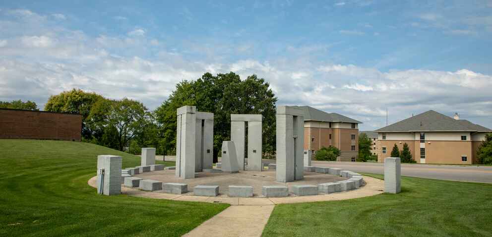 A stone circle monument with tall rectangular pillars and shorter blocks arranged in a circular formation on a grassy lawn, with buildings and trees in the background.
