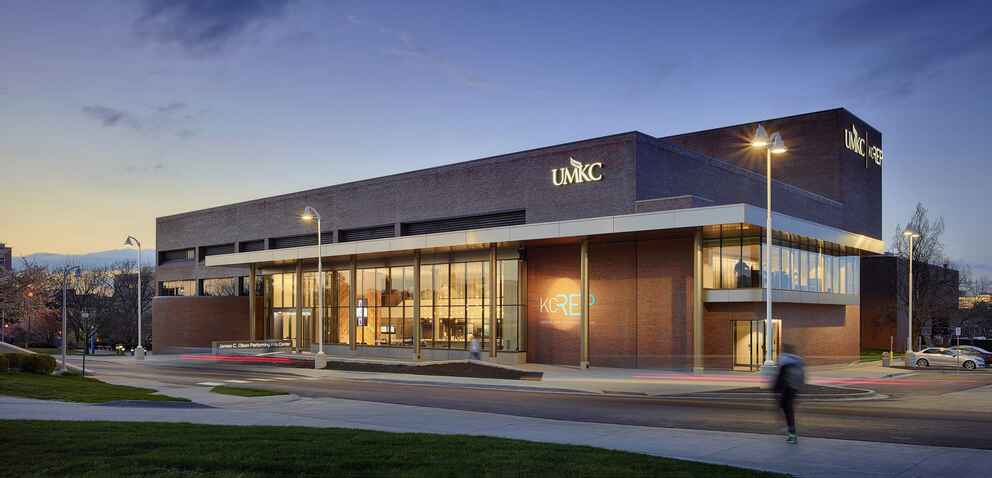The James C. Olson Performing Arts Center at UMKC, a modern building with large glass windows and brick walls, illuminated at dusk with a person walking outside.