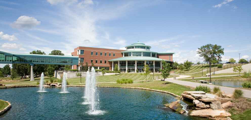 A scenic view of the UMSL campus featuring a modern brick building with large windows, a pond with fountains, and a pedestrian bridge under a clear blue sky.