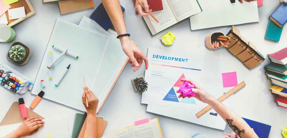 A group of people working together at a table filled with books, notebooks, markers, sticky notes, and a development project sheet, indicating a collaborative brainstorming session.